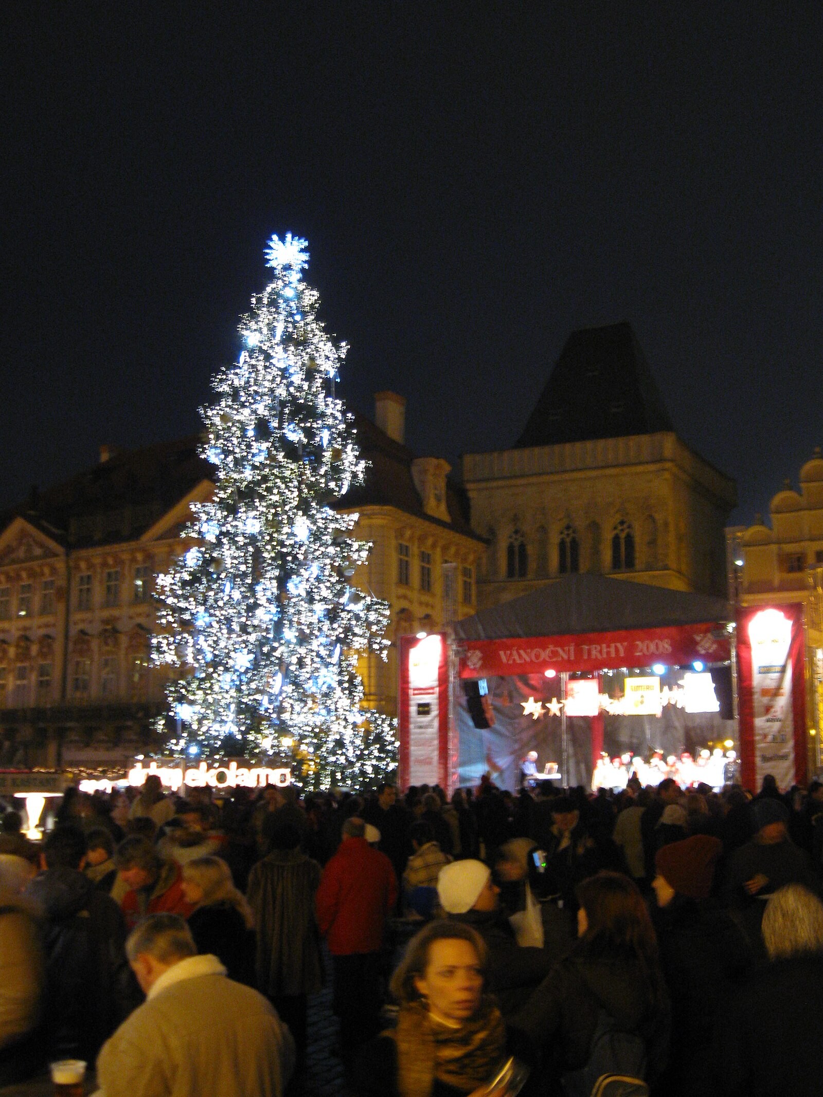 Old Town Square Christmas market at night with illuminated tree, stalls/crowd and evening programme.