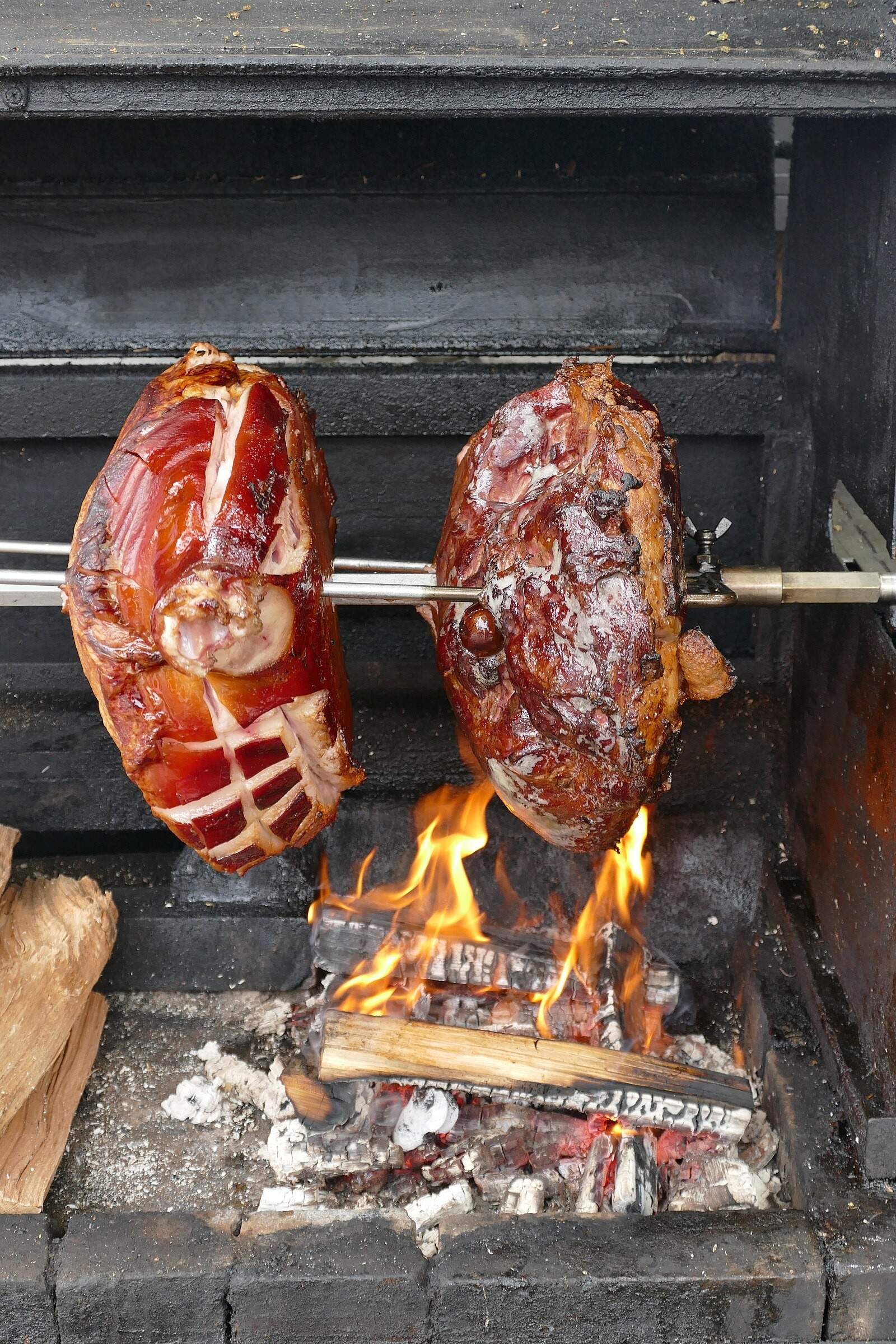 Large Prague hams roasting on spits over a wood fire at a food stall.