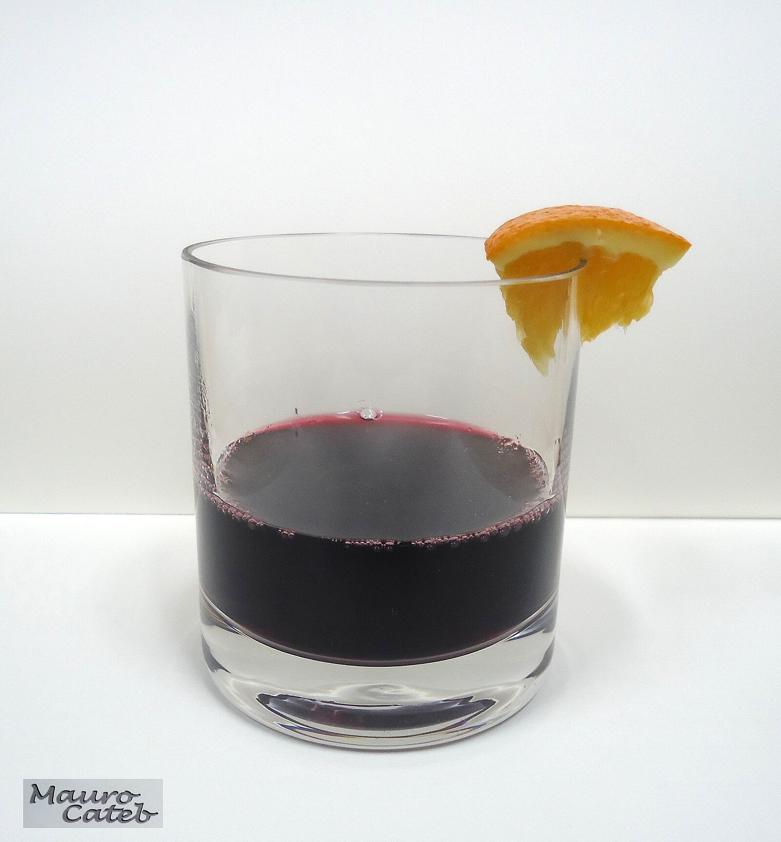 Glass of dark red mulled wine garnished with an orange wedge; small photographer signature is visible.