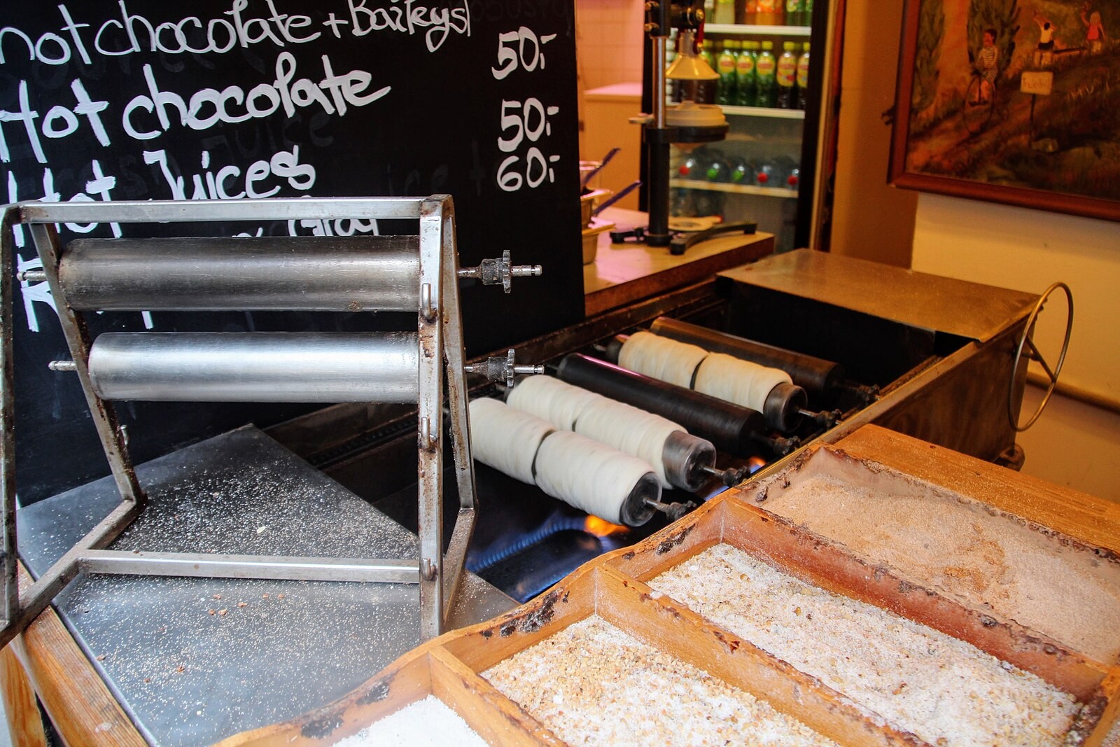 Trdelník cylinders rotating/baking over heat in a Prague shop, with sugar/coating preparation visible.
