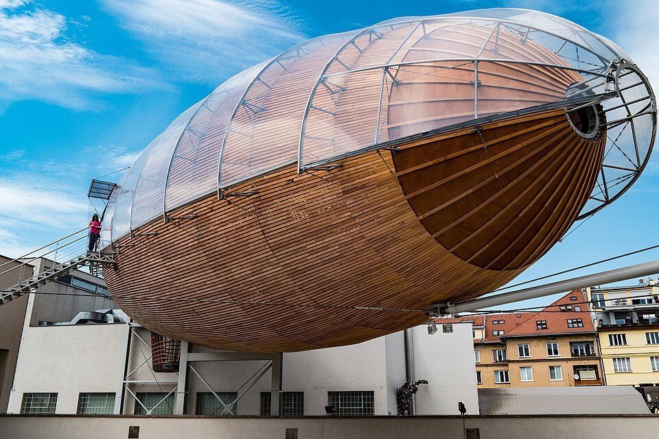 Large wooden-and-steel airship visibly mounted above a rooftop, seen from the bow.