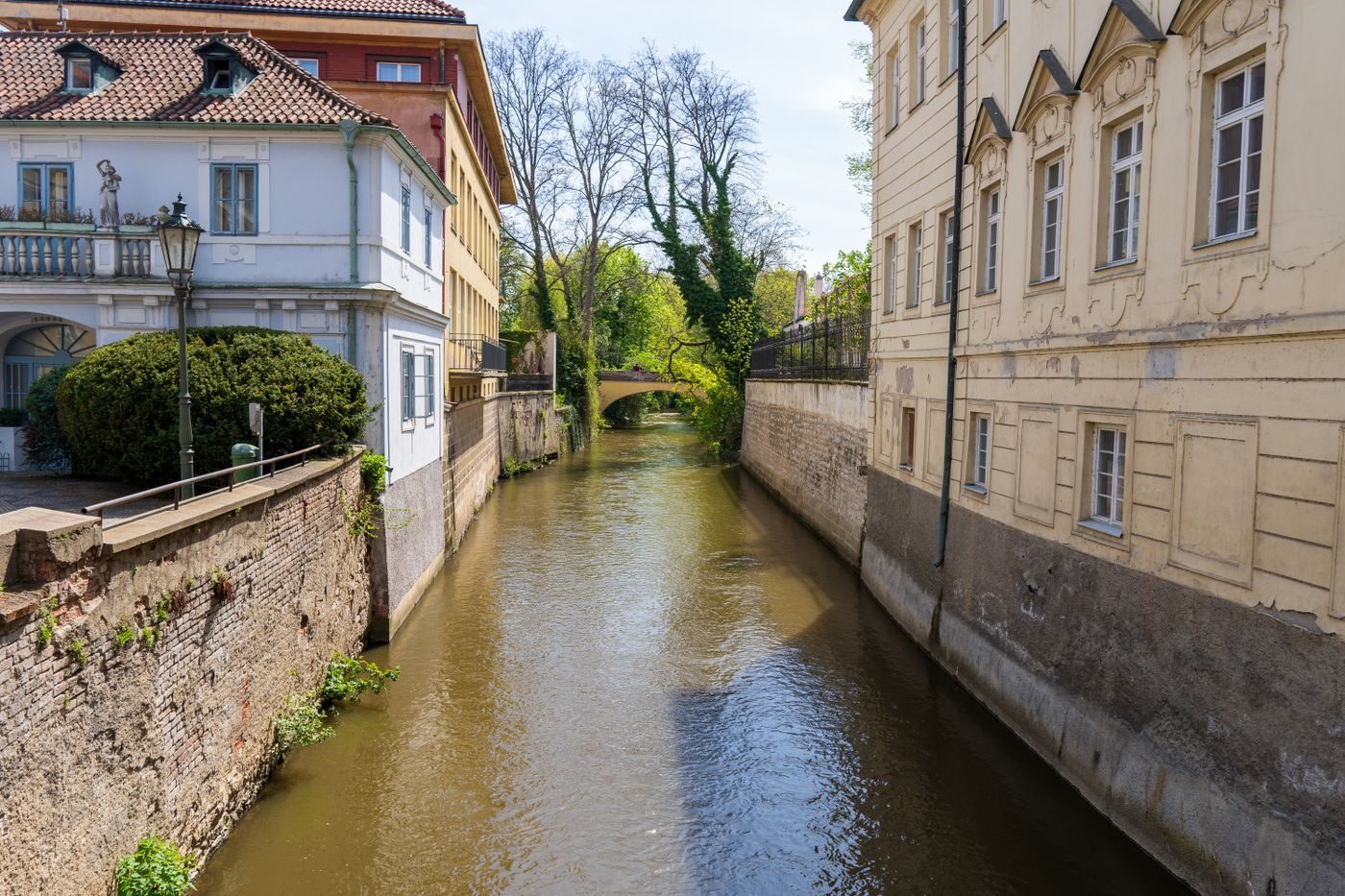 Narrow Prague waterway is visibly enclosed by walls/buildings, with a small bridge ahead.