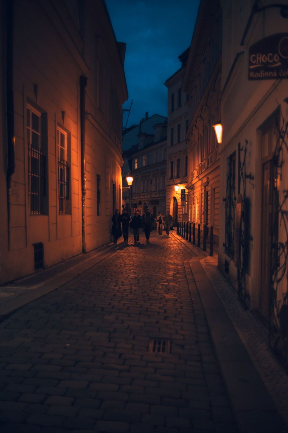 Pexels identifies a charming illuminated cobblestone street in Prague’s Old Town at night with passersby.
