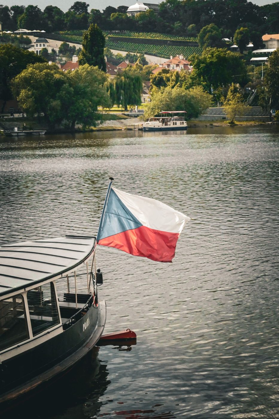 Foreground vessel carries a visible Czech flag with another passenger boat and Prague river context behind.