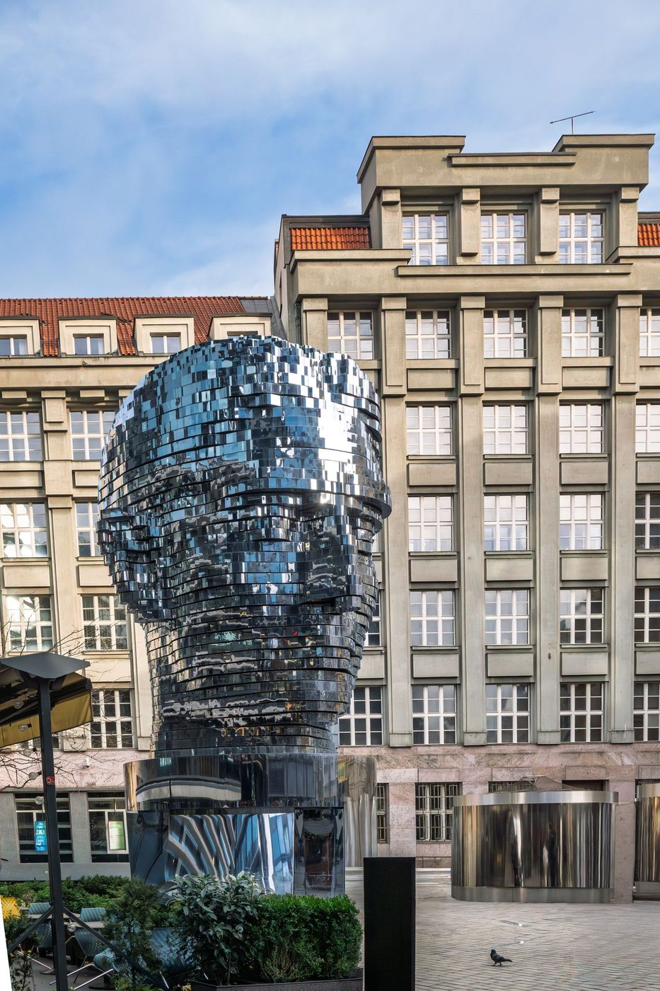Whole reflective Kafka head is visibly large against surrounding Prague architecture.