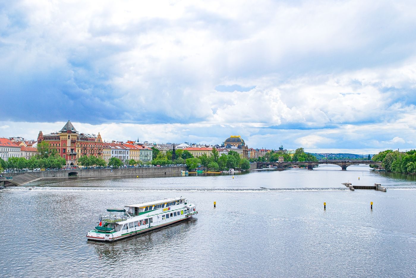 Low white/green passenger vessel is visibly traveling on the Vltava with Prague urban/bridge context.